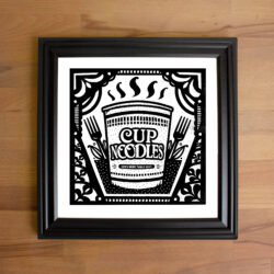 Print of a Cup Noodle illustration in a black frame on a wooden tabletop.